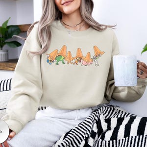 Op de afbeelding: Zandkleurige sweatshirt met een cartoonafbeelding van personages die oranje verkeerskegels dragen. Het ontwerp bevat een groene dinosaurus, een roze varken en andere figuren. De sweatshirt heeft een ronde hals.