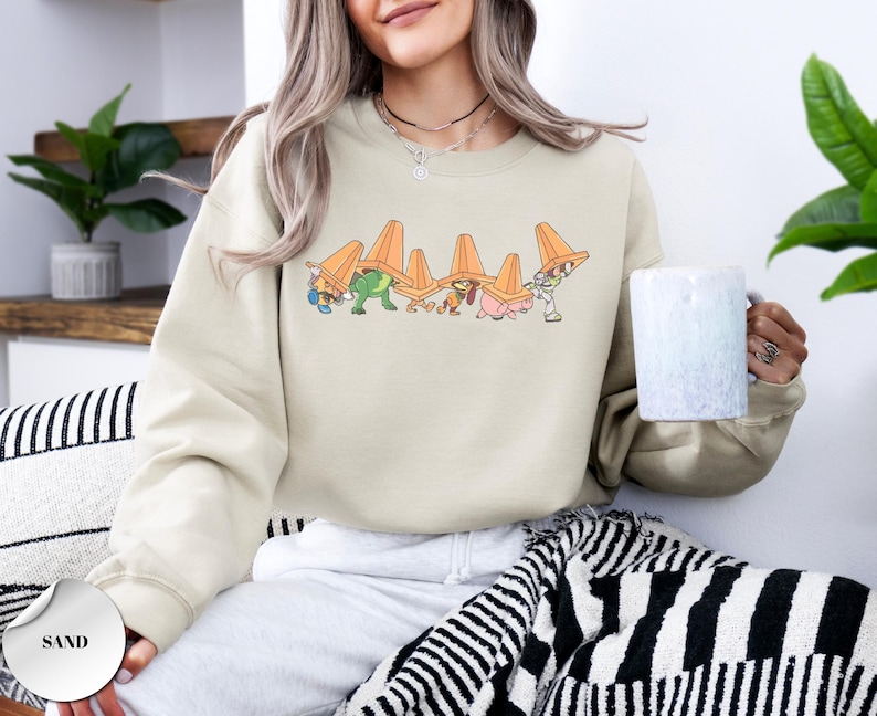 Op de afbeelding: Zandkleurige sweatshirt met een cartoonafbeelding van personages die oranje verkeerskegels dragen. Het ontwerp bevat een groene dinosaurus, een roze varken en andere figuren. De sweatshirt heeft een ronde hals.
