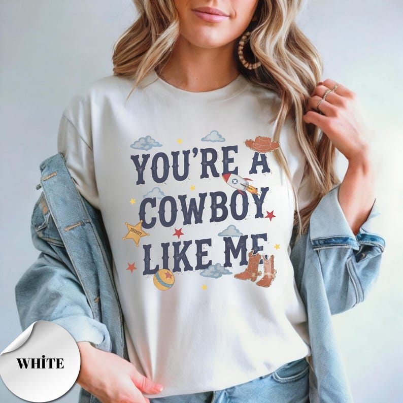 May include: A white short-sleeved t-shirt featuring the dark blue text "YOU'RE A COWBOY LIKE ME" surrounded by colorful graphic illustrations of a rocket, cowboy boots, a sheriff star, clouds, small stars, and a ball on the front chest area.