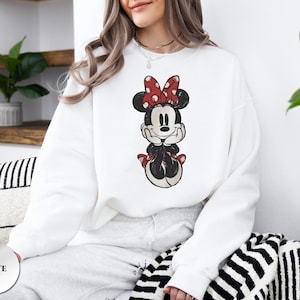 May include: White sweatshirt featuring a distressed graphic of Minnie Mouse. Minnie is depicted with a red bow with white polka dots. The word "WHITE" is printed on a white circle in the lower left corner.