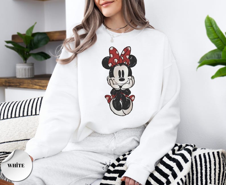 May include: White sweatshirt featuring a distressed graphic of Minnie Mouse. Minnie is depicted with a red bow with white polka dots. The word "WHITE" is printed on a white circle in the lower left corner.