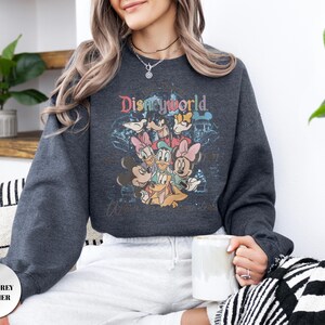 Op de afbeelding: Donkergrijze heather sweatshirt met een kleurrijke Disney World-afbeelding met Mickey Mouse, Minnie Mouse, Donald Duck en andere personages. De sweatshirt heeft een ronde hals en lange mouwen. De tekst "Disney World" staat bovenaan de afbeelding.