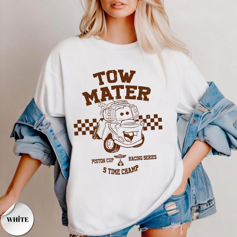 May include: A white t-shirt featuring a brown graphic of the character Tow Mater from the movie Cars. The design includes a checkered flag pattern, the text TOW MATER, PISTON CUP RACING SERIES, and S TIME CHAMP, all printed in a distressed brown font.