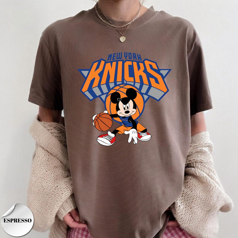 Puede incluir: Una camiseta marr&oacute;n de manga corta con un gr&aacute;fico de Mickey Mouse sosteniendo un bal&oacute;n de baloncesto. Sobre el personaje, el texto NEW YORK KNICKS aparece en letras naranjas con bordes azules sobre un elemento de fondo triangular azul.