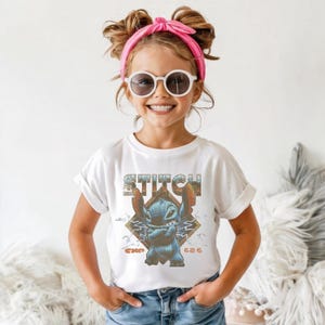 May include: A white short-sleeved t-shirt featuring a graphic of the blue character Stitch with arms crossed. The design includes the text STITCH in a retro font, the text EXP 626, and a diamond-shaped background with lightning bolt accents.
