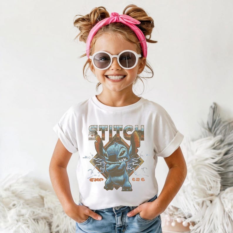 May include: A white short-sleeved t-shirt featuring a graphic of the blue character Stitch with arms crossed. The design includes the text STITCH in a retro font, the text EXP 626, and a diamond-shaped background with lightning bolt accents.