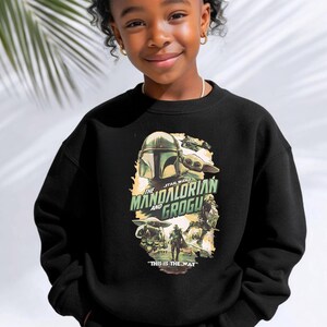 Puede incluir: Una sudadera negra de cuello redondo con un estampado gr&aacute;fico de The Mandalorian y Grogu. El dise&ntilde;o incluye el texto The Mandalorian and Grogu en letras verdes y la frase This is the way en blanco en la parte inferior, con varias ilustraciones.