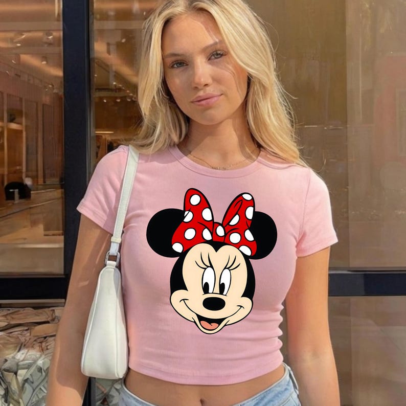 Puede incluir: Top corto rosa claro con un dise&ntilde;o de Minnie Mouse. El dise&ntilde;o muestra la cara de Minnie, un lazo rojo con lunares blancos y orejas negras. La parte superior tiene cuello redondo y mangas cortas.