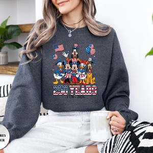 May include: A dark grey heather sweatshirt featuring a graphic of Disney characters including Mickey, Minnie, Donald, Daisy, Goofy, and Pluto wearing patriotic red, white, and blue attire. The text reads 250 YEARS 1776-2026 with American flag motifs.