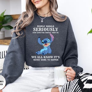 May include: A dark grey heather crewneck sweatshirt featuring a graphic of the blue character Stitch. The text reads: PEOPLE SHOULD SERIOUSLY STOP EXPECTING NORMAL FROM ME WE ALL KNOW IT'S NEVER GOING TO HAPPEN. The long-sleeved garment has ribbed cuffs.