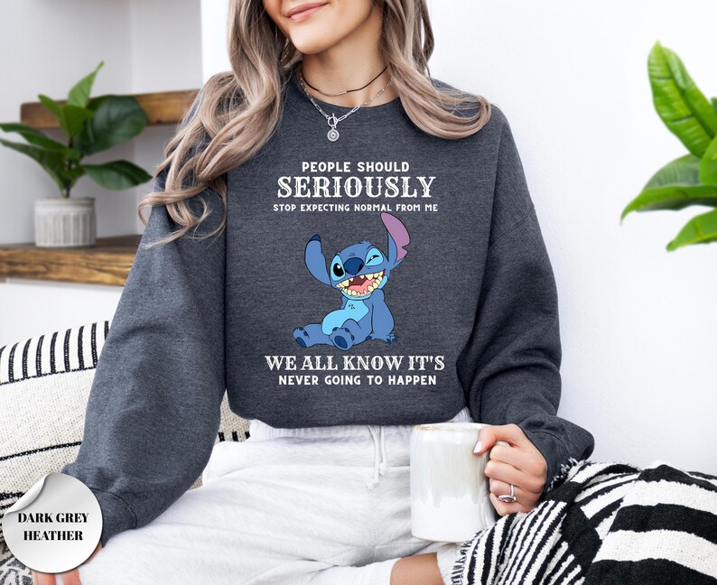 May include: A dark grey heather crewneck sweatshirt featuring a graphic of the blue character Stitch. The text reads: PEOPLE SHOULD SERIOUSLY STOP EXPECTING NORMAL FROM ME WE ALL KNOW IT'S NEVER GOING TO HAPPEN. The long-sleeved garment has ribbed cuffs.