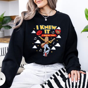 May include: A black long-sleeve sweatshirt featuring a graphic print of a person with long hair playing a guitar. The text reads I KNEW IT and I KNEW YOU, accompanied by a red hat, a red heart with TS, white clouds, musical notes, and yellow stars.