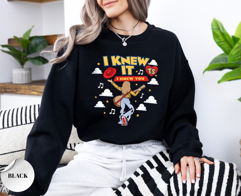 May include: A black long-sleeve sweatshirt featuring a graphic print of a person with long hair playing a guitar. The text reads I KNEW IT and I KNEW YOU, accompanied by a red hat, a red heart with TS, white clouds, musical notes, and yellow stars.
