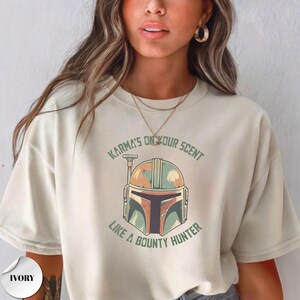 May include: An ivory short-sleeve t-shirt featuring a graphic of a stylized helmet in shades of green and tan. The text "KARMA'S ON YOUR SCENT" is printed in an arc above the helmet, and "LIKE A BOUNTY HUNTER" is printed in an arc below it.