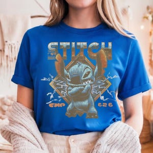 May include: A royal blue short-sleeve t-shirt featuring a graphic of the character Stitch with arms crossed. The design includes the text STITCH at the top, EXP 626 at the bottom, and a diamond-shaped background with lightning bolt accents.