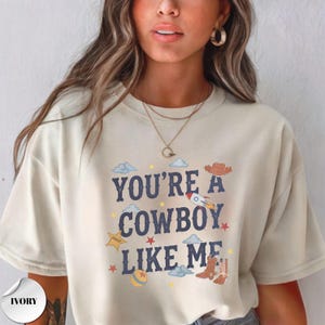 May include: An ivory short-sleeved t-shirt featuring the dark blue text "YOU'RE A COWBOY LIKE ME" centered on the chest. The design includes graphic illustrations of a brown cowboy hat, a pair of brown cowboy boots, a rocket, clouds, and stars.