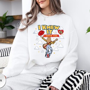 May include: A white long-sleeve sweatshirt featuring a graphic print with the text I KNEW IT I KNEW YOU TS. The design includes a cartoon figure playing a guitar, a red hat, a red heart, musical notes, and white clouds on a white background.