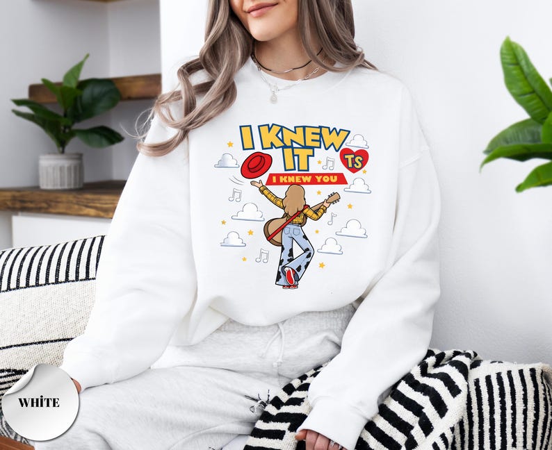 May include: A white long-sleeve sweatshirt featuring a graphic print with the text I KNEW IT I KNEW YOU TS. The design includes a cartoon figure playing a guitar, a red hat, a red heart, musical notes, and white clouds on a white background.