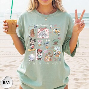 K&ouml;nnte beinhalten: Hellgr&uuml;nes T-Shirt mit verschiedenen Disney-Figuren und Wahrzeichen. Das Shirt zeigt Micky Maus, Minnie Maus, Donald Duck und Goofy sowie den Text "Walt Disney World" und "2026". Der Satz "The Most Magical Place on Earth" ist ebenfalls auf dem Shirt aufgedruckt.