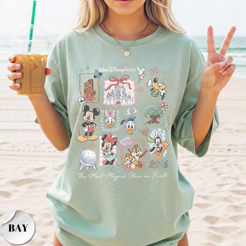 K&ouml;nnte beinhalten: Hellgr&uuml;nes T-Shirt mit verschiedenen Disney-Figuren und Wahrzeichen. Das Shirt zeigt Micky Maus, Minnie Maus, Donald Duck und Goofy sowie den Text "Walt Disney World" und "2026". Der Satz "The Most Magical Place on Earth" ist ebenfalls auf dem Shirt aufgedruckt.