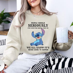 May include: A sand-colored crewneck sweatshirt featuring a graphic of the blue character Stitch. The text reads: PEOPLE SHOULD SERIOUSLY STOP EXPECTING NORMAL FROM ME WE ALL KNOW IT'S NEVER GOING TO HAPPEN. The sweatshirt has long sleeves and a ribbed collar.