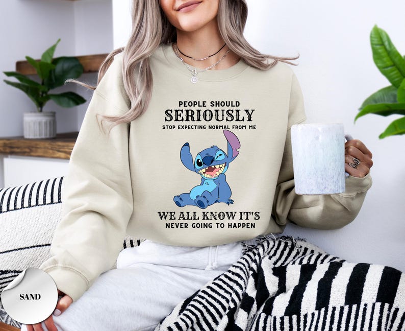 May include: A sand-colored crewneck sweatshirt featuring a graphic of the blue character Stitch. The text reads: PEOPLE SHOULD SERIOUSLY STOP EXPECTING NORMAL FROM ME WE ALL KNOW IT'S NEVER GOING TO HAPPEN. The sweatshirt has long sleeves and a ribbed collar.