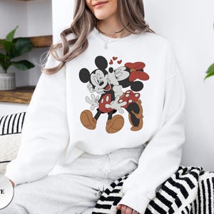 May include: White sweatshirt featuring a cartoon illustration of Mickey Mouse and Minnie Mouse kissing, with hearts above them. Mickey is wearing red shorts and Minnie has a red bow. The word "WHITE" is printed on a white circle.