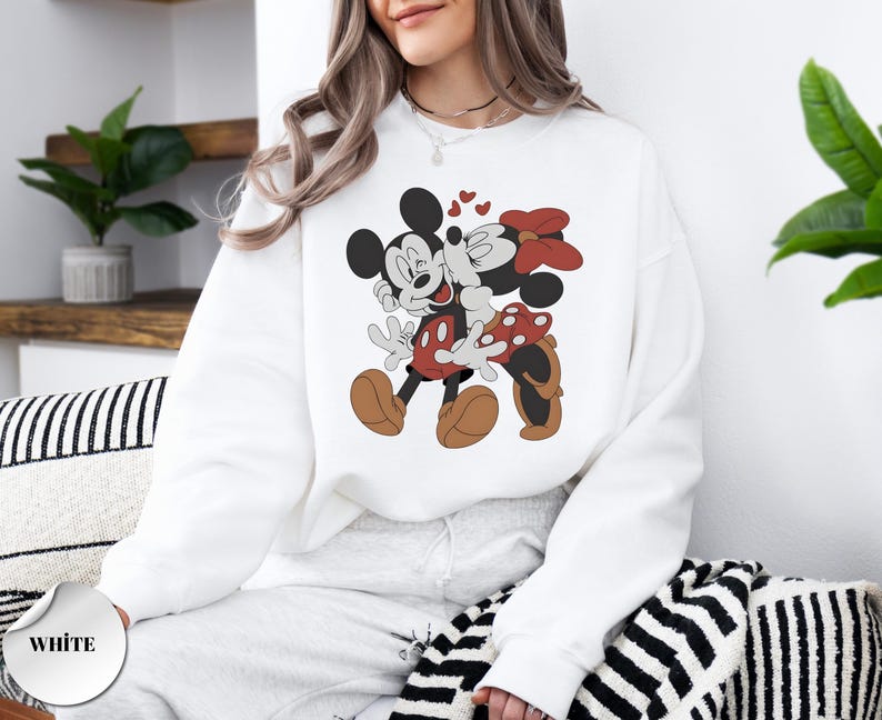 May include: White sweatshirt featuring a cartoon illustration of Mickey Mouse and Minnie Mouse kissing, with hearts above them. Mickey is wearing red shorts and Minnie has a red bow. The word "WHITE" is printed on a white circle.