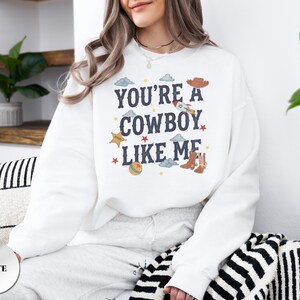May include: A white long-sleeve crewneck sweatshirt featuring the dark blue text "YOU'RE A COWBOY LIKE ME" centered on the chest. The design includes small colorful illustrations of a rocket, cowboy boots, a hat, a ball, stars, and clouds.