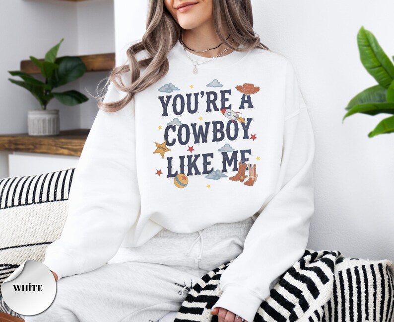 May include: A white long-sleeve crewneck sweatshirt featuring the dark blue text "YOU'RE A COWBOY LIKE ME" centered on the chest. The design includes small colorful illustrations of a rocket, cowboy boots, a hat, a ball, stars, and clouds.