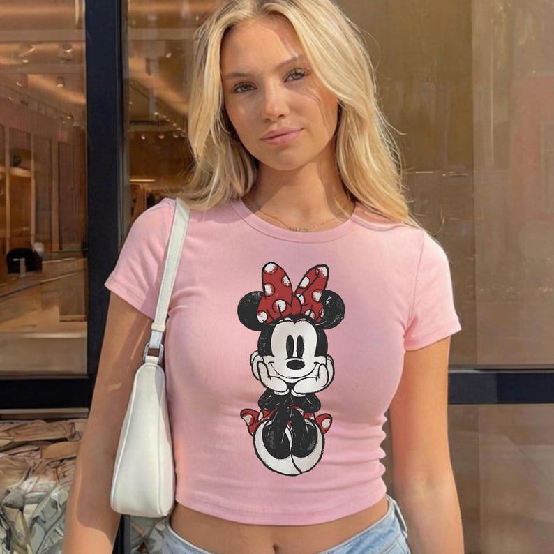 May include: A light pink crop top featuring a Minnie Mouse graphic. The graphic shows Minnie Mouse in black and white with a red polka dot bow. The top has short sleeves and a round neckline. A white purse is hanging from the shoulder.