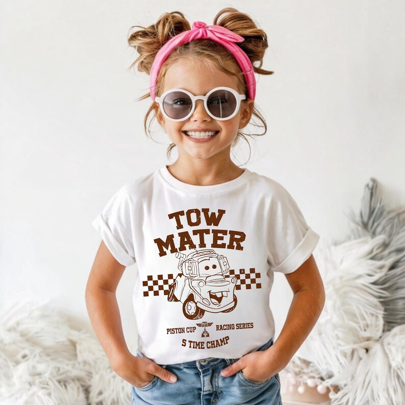 May include: A white short-sleeve t-shirt featuring a brown graphic of the character Tow Mater. The design includes the text "TOW MATER", a checkered pattern, and the words "PISTON CUP RACING SERIES" and "S TIME CHAMP" printed in brown ink.