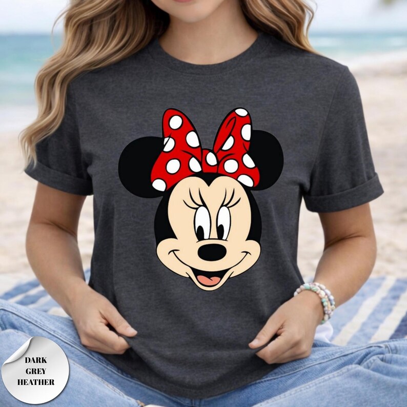 Puede incluir: Camiseta gris oscuro jaspeado con un gr&aacute;fico de Minnie Mouse. El gr&aacute;fico muestra la cara de Minnie con un lazo rojo con lunares blancos. El texto "DARK GREY HEATHER" est&aacute; en una pegatina en la esquina inferior izquierda.