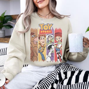 K&ouml;nnte beinhalten: Ein sandfarbenes Sweatshirt mit Rundhalsausschnitt und einem Grafikdruck der Toy Story-Figuren Bullseye, Woody, Buzz Lightyear und Jessie. Das Design enth&auml;lt das Toy Story-Logo in gelber und roter Schrift &uuml;ber den Charakter-Illustrationen.