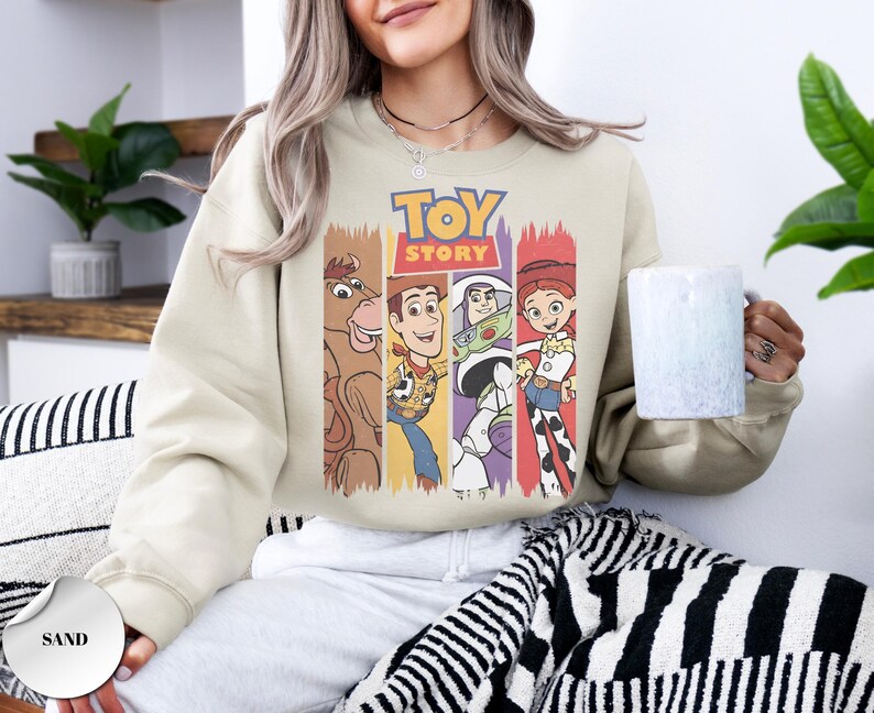 K&ouml;nnte beinhalten: Ein sandfarbenes Sweatshirt mit Rundhalsausschnitt und einem Grafikdruck der Toy Story-Figuren Bullseye, Woody, Buzz Lightyear und Jessie. Das Design enth&auml;lt das Toy Story-Logo in gelber und roter Schrift &uuml;ber den Charakter-Illustrationen.
