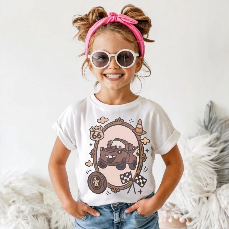 May include: A white short-sleeve t-shirt featuring a brown cartoon tow truck graphic inside an oval frame. The design includes a Route 66 sign, a traffic cone, a tire, and checkered flags. The shirt is paired with blue denim jeans.