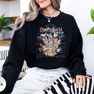 Op de afbeelding: Zwart sweatshirt met een kleurrijke afbeelding van Disney-personages, waaronder Mickey en Minnie Mouse, Donald en Daisy Duck en Goofy. Het ontwerp bevat de woorden "Walt Disney World" en "Where dreams come true."