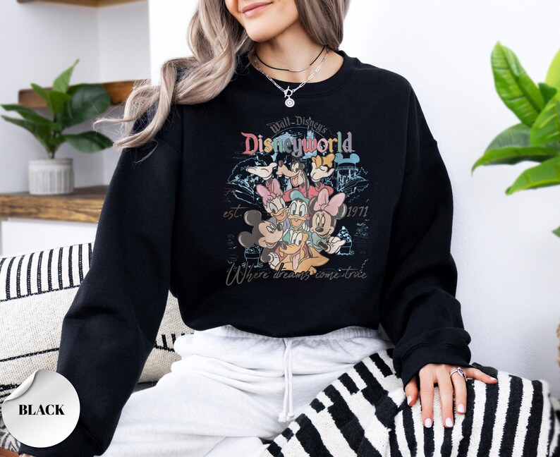 Op de afbeelding: Zwart sweatshirt met een kleurrijke afbeelding van Disney-personages, waaronder Mickey en Minnie Mouse, Donald en Daisy Duck en Goofy. Het ontwerp bevat de woorden "Walt Disney World" en "Where dreams come true."