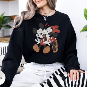 May include: Black sweatshirt featuring a graphic of Mickey and Minnie Mouse embracing, with hearts above them. Minnie wears a red bow and polka-dot skirt. The sweatshirt has a round neckline and long sleeves. The word "BLACK" is printed on a white sticker.