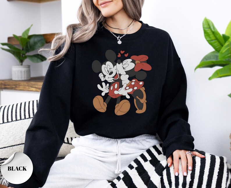 May include: Black sweatshirt featuring a graphic of Mickey and Minnie Mouse embracing, with hearts above them. Minnie wears a red bow and polka-dot skirt. The sweatshirt has a round neckline and long sleeves. The word "BLACK" is printed on a white sticker.