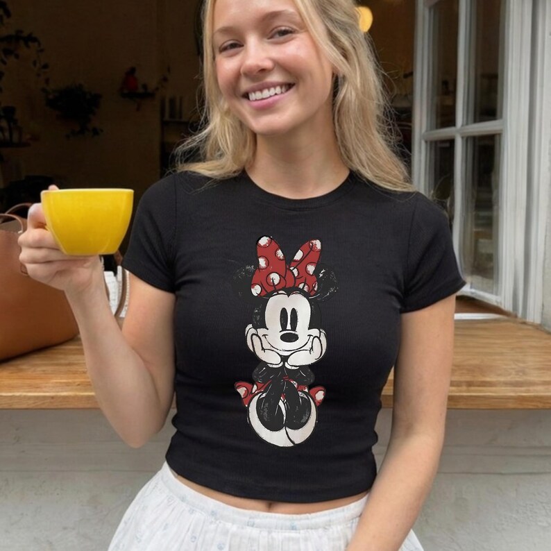 May include: Black crop top featuring a Minnie Mouse graphic. The graphic shows Minnie Mouse with a red and white polka dot bow. The shirt is short-sleeved and has a round neckline. The shirt is worn with a white skirt.