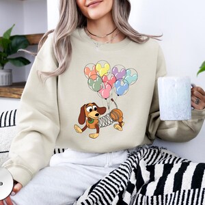 May include: Sand-colored sweatshirt featuring a cartoon illustration of Slinky Dog holding colorful Mickey Mouse-shaped balloons. The design includes brown, yellow, pink, and blue balloons. The sweatshirt is a neutral color.
