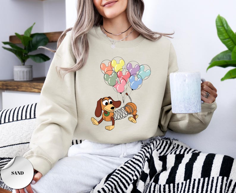 May include: Sand-colored sweatshirt featuring a cartoon illustration of Slinky Dog holding colorful Mickey Mouse-shaped balloons. The design includes brown, yellow, pink, and blue balloons. The sweatshirt is a neutral color.