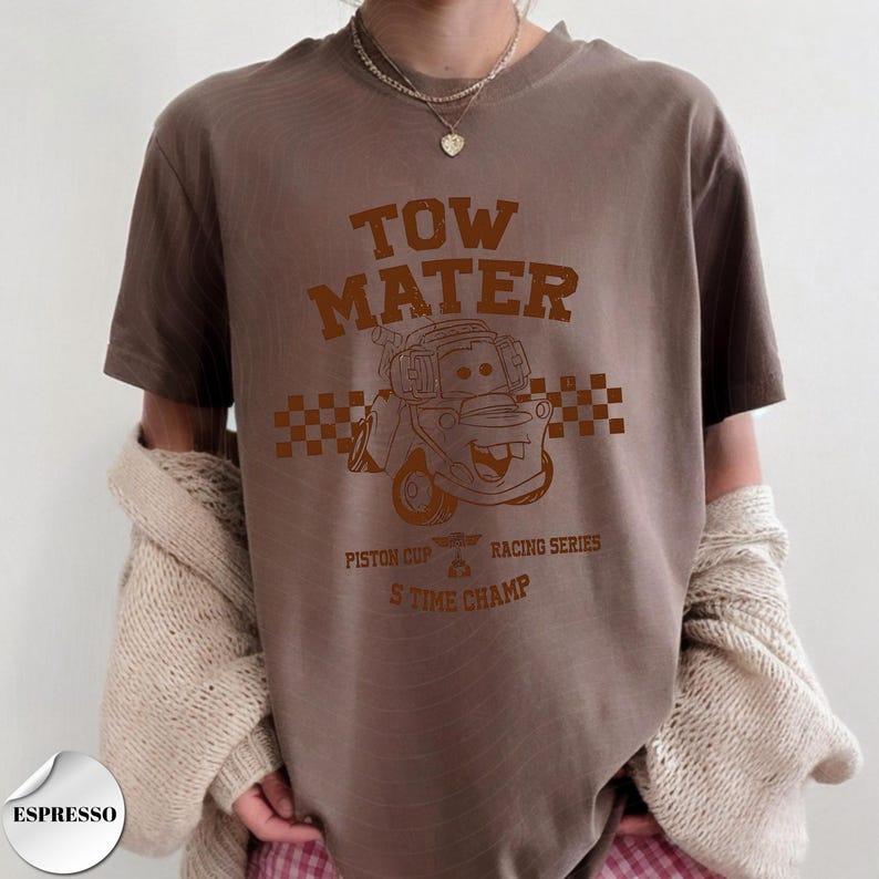May include: A brown short-sleeve t-shirt featuring a dark brown graphic of the character Tow Mater. The design includes a checkered pattern, the text TOW MATER, PISTON CUP RACING SERIES, and 5 TIME CHAMP. A small circular label in the corner reads ESPRESSO.