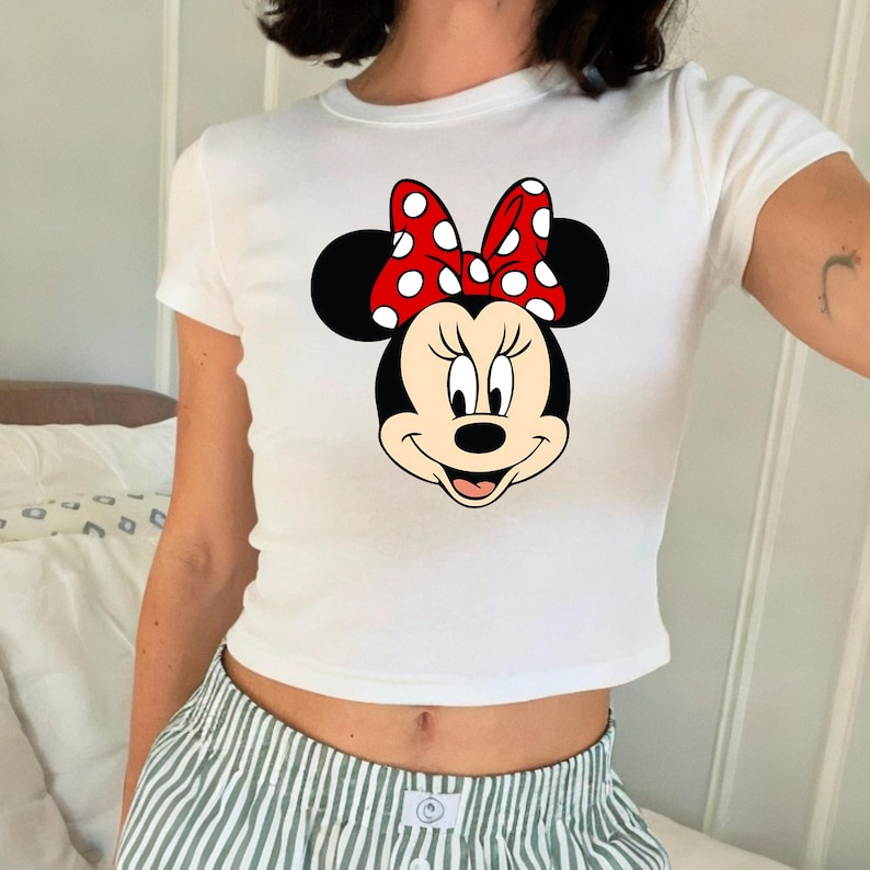Puede incluir: Top corto blanco con un gr&aacute;fico de Minnie Mouse. El gr&aacute;fico muestra la cara de Minnie con un lazo rojo con lunares blancos. La camiseta tiene mangas cortas y cuello redondo.