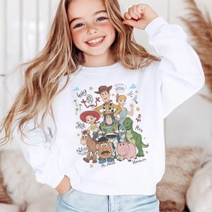 May include: A white long-sleeve sweatshirt featuring a colorful graphic print of Toy Story characters including Woody, Buzz Lightyear, Jessie, Rex, Hamm, Mr. Potato Head, Bullseye, Forky, and Bo Peep, with their names written in a cursive script.