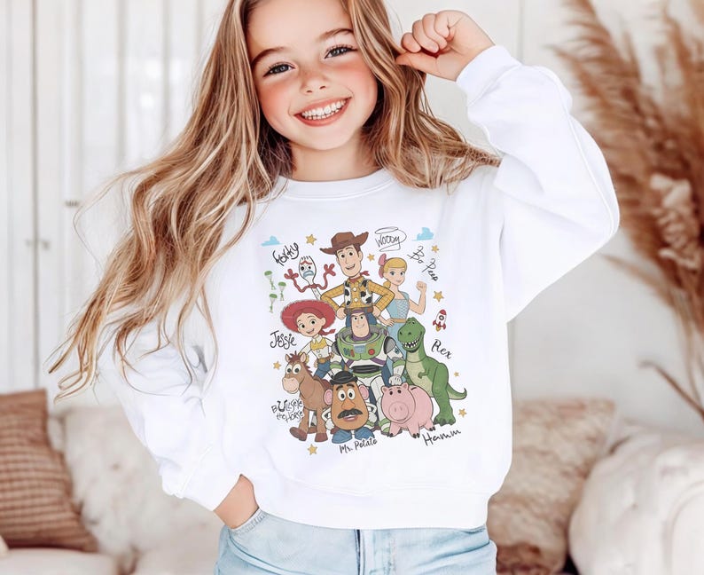 May include: A white long-sleeve sweatshirt featuring a colorful graphic print of Toy Story characters including Woody, Buzz Lightyear, Jessie, Rex, Hamm, Mr. Potato Head, Bullseye, Forky, and Bo Peep, with their names written in a cursive script.