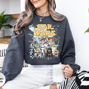 Puede incluir: Una sudadera de cuello redondo en color gris jaspeado oscuro con un estampado gr&aacute;fico de personajes de Star Wars reimaginados como figuras de Toy Story. El dise&ntilde;o incluye el logotipo amarillo de Star Wars sobre ilustraciones de Woody, Jessie y otros.