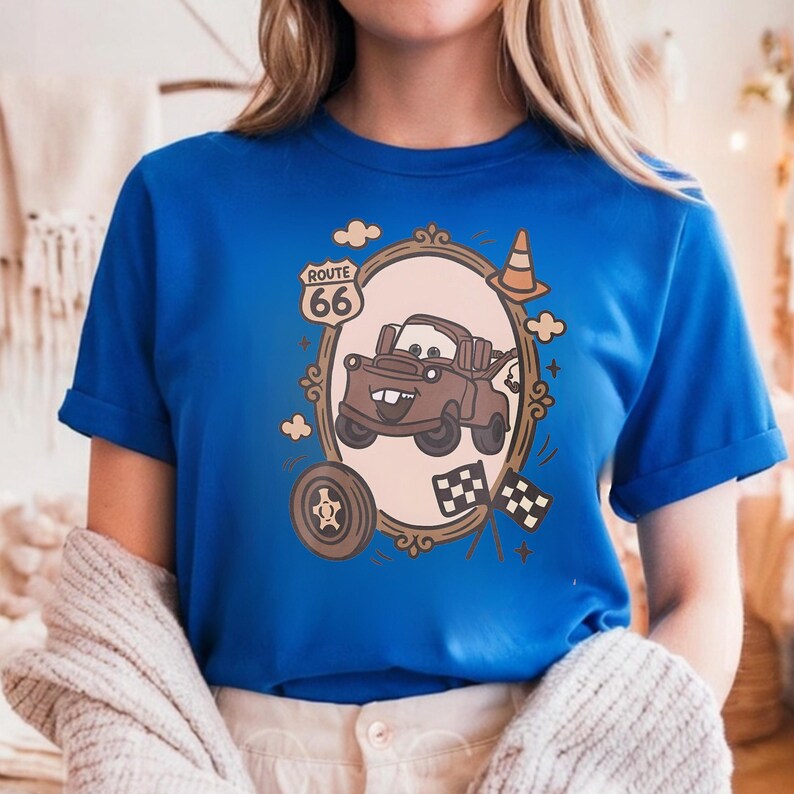 May include: A bright blue t-shirt featuring a graphic print of a brown tow truck character inside an ornate oval frame. The design includes a Route 66 sign, a traffic cone, a tire, and two checkered racing flags in shades of brown and cream.