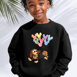 May include: Black sweatshirt featuring a cartoon image of Slinky Dog from Toy Story. The design includes colorful Mickey Mouse-shaped balloons. The sweatshirt is a crew neck style.